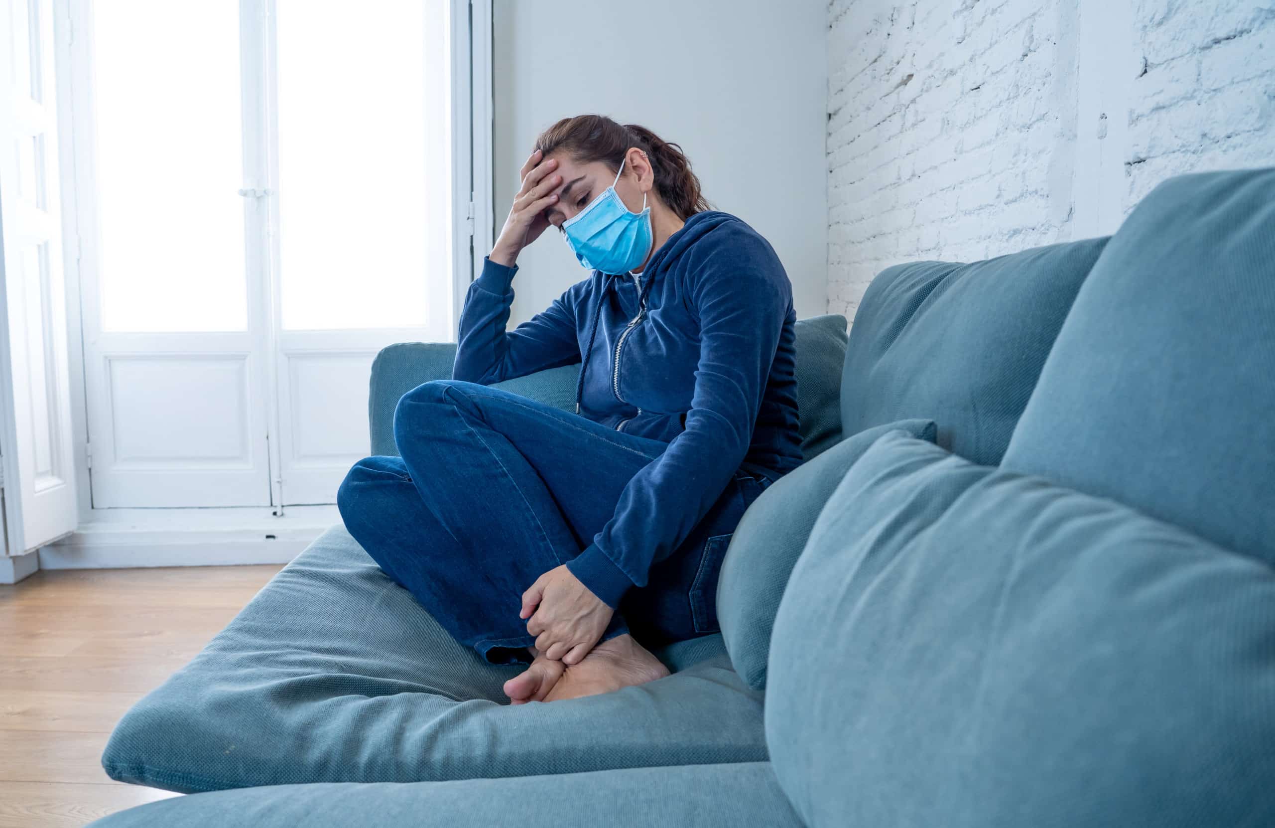 Sad woman sitting on the sofa with protective mask at home.