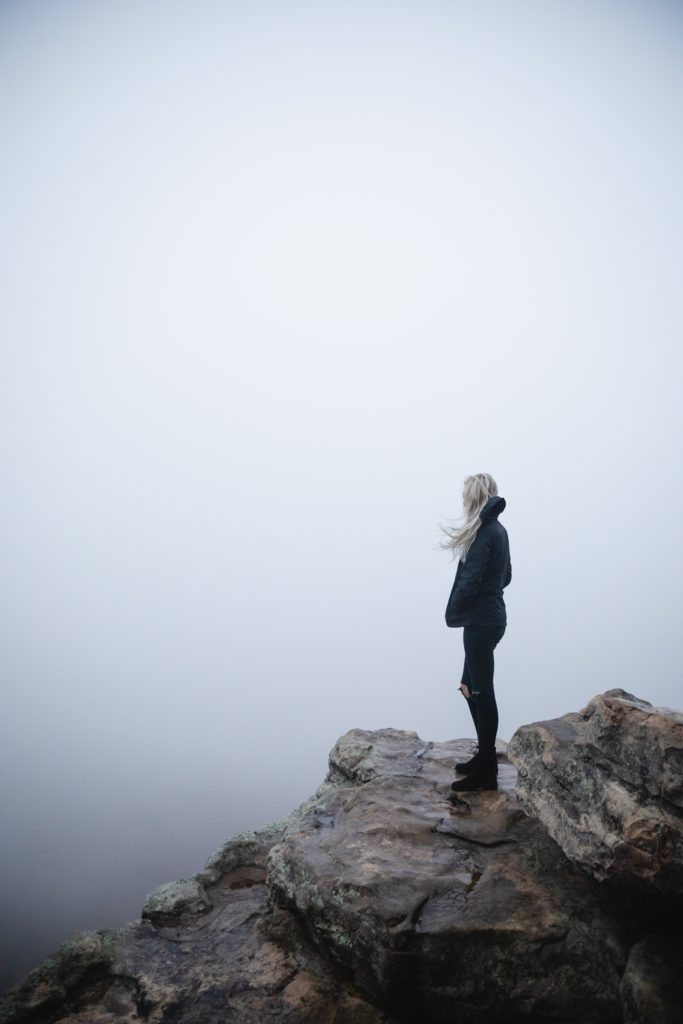 Woman standing on the cliff looking at down at the clouds hanging near her.