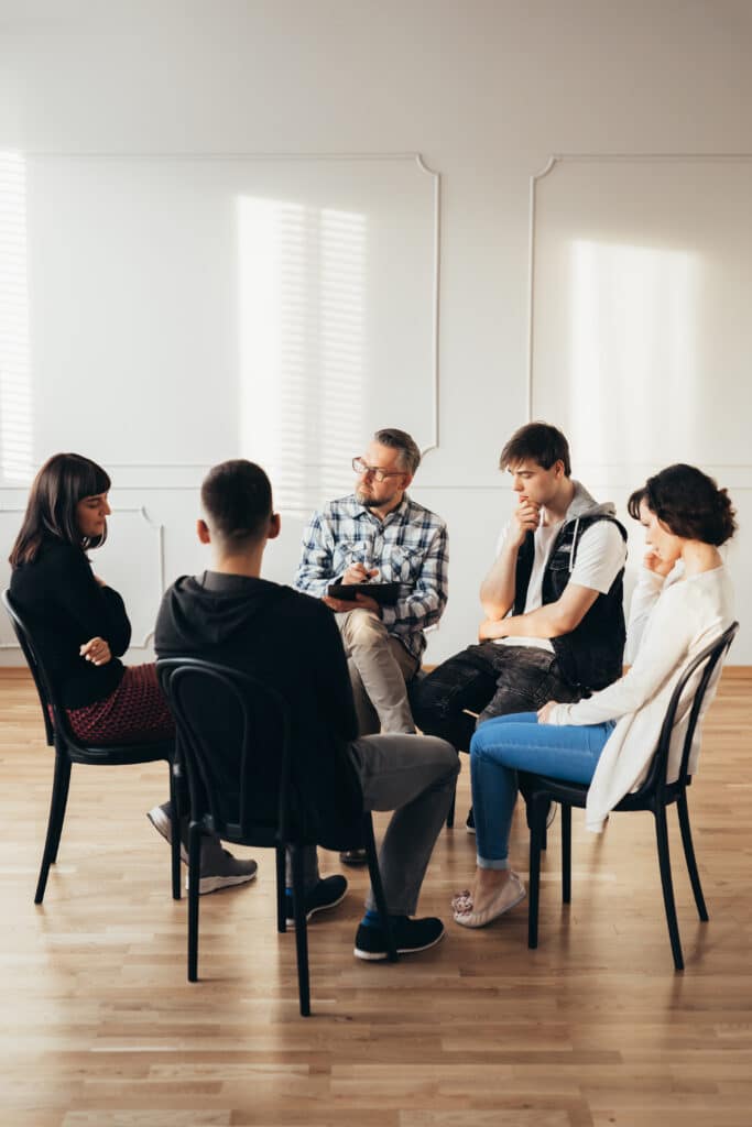 Group Of People Of Different Ages Sits In A= Circle for a Group Therapy Session with Therapist.