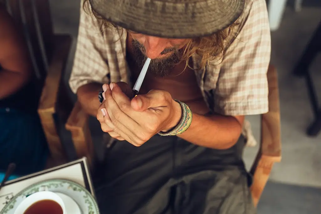 man lighting marijuana cigarette while sitting in a chair wearing rounf hat